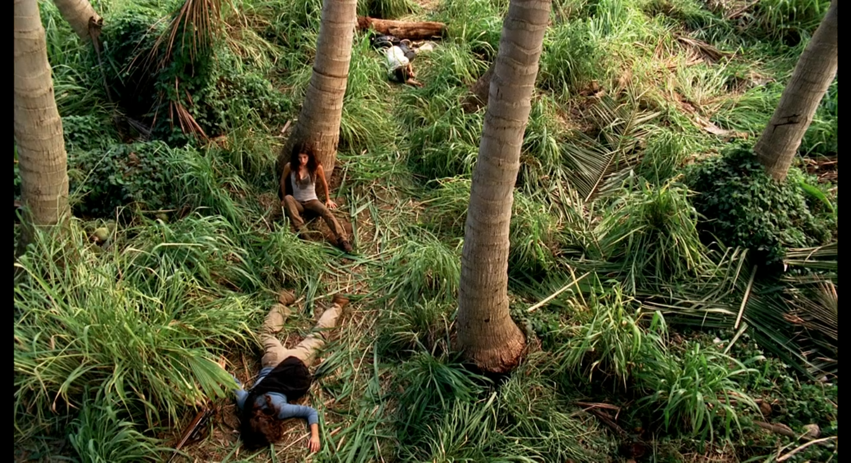 High angle. Alex Rousseau sits against a tree. The corpses of Karl and Rousseau lie on the ground.