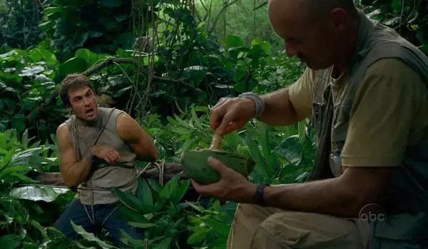 Boone Carlyle (Ian Somerhalder) crouches in a jungle, securely bound. He remonstrates with John Locke (Terry O’Quinn), seen in the foreground of the shot, grinding something in a bowl with a pestle.