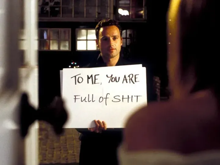 A man holds cards with hand written messages. The message on the card he's holding says "TO ME YOU ARE FULL OF SHIT